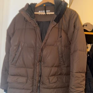 Brun dunjacka från Everest med huva - Säljer en brun dunjacka från Everest i storlek 38. Jackan har en skön huva, dragkedja och stora fickor framtill. Yttermaterialet är nylon och fyllningen består av 50% dun och 50% fjäder. Perfekt för kalla dagar när du vill hålla värmen.