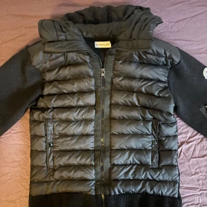 Svart Moncler cardigan med huva - Svart cardigan från Moncler med stickade ärmar och quiltad framsida. Jackan har en stor vadderad huva, dragkedja framtill och fickor med knappar. Ärmarna är ribbstickade och har en liten ficka med dragkedja samt Moncler-logga på ena ärmen. Använt en gång. Kan ta bort alla fjädrar. Lägg gärna ett prisförslag