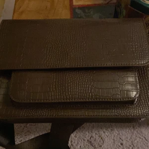 Brun axelväska med krokopräglat mönster - Snygg brun axelväska med krokopräglat mönster och bred justerbar axelrem. Väskan har ett stilrent, rektangulärt format och flera fack framtill. Materialet är skinnimitation med struktur som ger en cool vibe. Perfekt för dig som gillar detaljer och vill sticka ut.