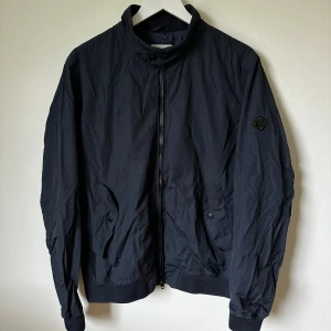 Mörkblå vindjacka från J.Lindeberg - Snygg mörkblå vindjacka från J.Lindeberg med hög krage och dragkedja framtill. Jackan har ribbade muddar, två fickor med knappstängning och diskret logga på ärmen. Perfekt för dig som gillar stilren och sportig look. 1F1