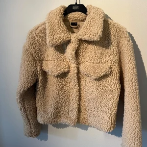 Beige teddyjacka från Gina Tricot XS - Supermjuk och fluffig teddyjacka från Gina Tricot i beige. Jackan har stora bröstfickor med lock, klassisk krage och stängs framtill. Perfekt för kyliga dagar och ger en riktigt cozy vibe. Passar dig som gillar en trendig och avslappnad stil.