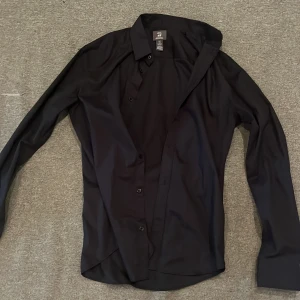 Svart slim fit skjorta från H&M - Svart skjorta från H&M i slim fit-modell med klassisk krage och långa ärmar. Skjortan har knappar framtill och är tillverkad i ett lättskött material som är enkel att stryka. Perfekt för dig som gillar en stilren och clean look.
