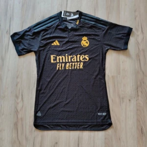 Svart Real Madrid fotbollströja Adidas - Snygg svart Real Madrid fotbollströja från Adidas med gula detaljer och klubbmärke på bröstet. Tröjan har korta ärmar, klassisk krage och är tillverkad i ett lätt, återvunnet funktionsmaterial med HEAT.RDY-teknologi. Perfekt för dig som älskar fotboll och vill sticka ut på planen.