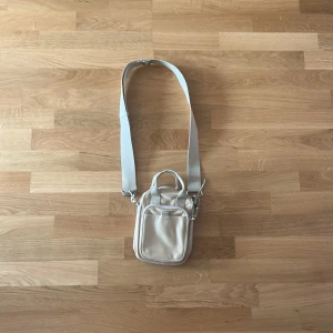 Beige axelväska från H&M - Säljer en stilren beige axelväska från H&M med justerbar bred rem och praktiskt handtag. Väskan har ett stort huvudfack, en mindre ficka framtill med dragkedja och silvriga detaljer. Perfekt storlek för mobil, plånbok och andra essentials.💕💕