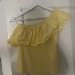 Gul offshoulder topp med volang - Söt gul offshoulder topp i stl 146/152 som passar som en xs med bred volang längs överdelen. Perfekt för varma dagar och ger en somrig vibe. Tunn och luftig, passar till både jeans och kjol. Enkel och stilren design.