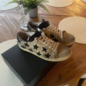 Saint Laurent sneakers med stjärnor - Beiga Saint Laurent sneakers i mocka med svarta stjärnor på sidorna och svarta detaljer vid hälen. Skorna har vit snörning och en klassisk rund tå. Perfekt för dig som vill sticka ut med en cool och unik look. Defekter som ni ser på bilderna. Box kommer inte med. Hör av er vid fler frågor. Skomakare!!