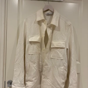 Beige overshirt från Zara - Stilren beige overshirt från Zara med klassisk krage och stora bröstfickor. Jackan har knäppning framtill och raka ärmar. Perfekt att slänga över en hoodie eller t-shirt för en clean look.
