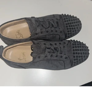 Christian Louboutin svarta sneakers med nitar - Grymma gråa sneakers från Christian Louboutin i mocka med coola svarta nitar på tån och klassisk röd sula. Skorna har snörning och en rund tå, perfekt för dig som vill sticka ut med en lyxig och edgy stil.