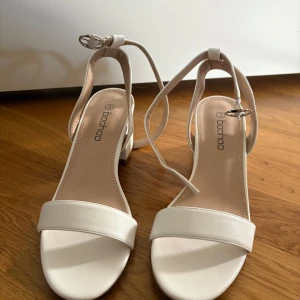 Vita sandaler med klack från Boohoo - Stilrena vita sandaler från Boohoo med bred rem över tårna och justerbar rem runt ankeln. Skorna har en chunky låg klack och öppen tå, perfekta för sommarens alla tillfällen. Tillverkade i syntetmaterial med mjuk innersula för extra komfort.