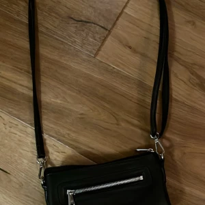 Svart axelväska med dragkedja😊 - Snygg svart axelväska med lång rem och silverfärgad dragkedja framtill. Väskan har en stilren och enkel design som passar till många olika outfits. Perfekt storlek för att få med det viktigaste och lätt att bära över axeln.