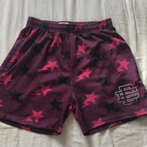 Lila meshshorts med stjärnmönster - Snygga lila meshshorts med elastisk midja och coolt stjärnmönster i rosa och svart. Perfekt för dig som gillar sportig stil och vill sticka ut med färg och mönster. Shortsen har en luftig känsla och ett tryckt emblem på ena benet. Märket är Eric Emanuel, dom är väldigt små i storleken, det står L men passar M/S