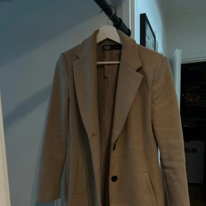 Beige kappa från Zara, stl M - OV:100€ Stilren beige kappa från Zara i klassisk rak modell med snygg krage och knäppning framtill. Långa ärmar, två fickor och slits bak. Perfekt för dig som gillar en clean och tidlös look. Fodrad insida för extra komfort. 