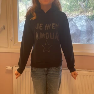 Svart hoodie med tryck från Gina Tricot - Svart hoodie från Gina Tricot med texten 'JE M'EN AMOUR' och en stjärna framtill. Skriv om ni har några fler frågor eller vill ha fler bilder🫶🏼💕