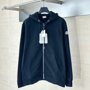 Svart Moncler hoodie med dragkedja - Svart hoodie från Moncler med vit logga på huvan och patch på ärmen. Tröjan har dragkedja framtill, snörning i huvan och känguruficka. Tillverkad i mjuk bomull, perfekt för en chill och stilren look.