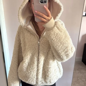 Beige teddyjacka med huva från Zara - Kikar intresse för min supermysiga teddyjacka från Zara i beige med dragkedja och stor huva. Jackan har ribbade muddar och en oversized passform som ger en chill vibe. Perfekt för kalla dagar när du vill vara både varm och snygg.