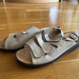 Beige sandaler  - Beiga sandaler i skinn från Cinnamon med justerbara remmar och broderade blommor i rött och blått på ovandelen. Skorna har öppen tå, platt sula och en bekväm passform, perfekta för varma dagar. Högra skorn har några svarta färg fläckar.
