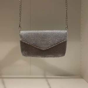 Glittrig silver kuvertväska från Glitter - Säljer en glittrig kuvertväska från Glitter i silver med kedjerem. Väskan är täckt av små glittrande stenar och har ett innerfoder i silkesliknande material. Väskan är bara använd en gång. 🎀💗👛