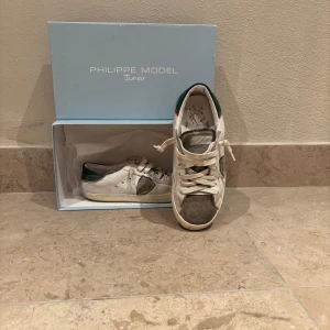 Philippe Model Junior vita sneakers MED LÅDA - Snygga Philippe Model Junior sneakers i vitt skinn med grå mockadetaljer på tå och sidor samt grön häl. Klassisk låg modell med snörning och platt sula. Perfekta för dig som gillar italiensk design och streetstyle. Kommer med låda som är i bra skick. Stl 37-38 (min storlek) Riktigt snygga och grisch och perfekta till slutet av sommaren och hösten !hälen ör väldigt sliten! Men fortfarande fungerande och intakt
