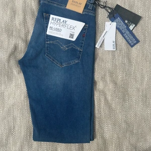 Replay Hyperflex Grover jeans blå - Blå Replay Hyperflex Grover jeans med straight fit. Klassisk femficksmodell med snygga kontrastsömmar och diskret röd detalj på bakfickan. Tillverkade i återvunnen bomull och polyester för extra stretch och hållbarhet. Perfekta för en clean och avslappnad look.