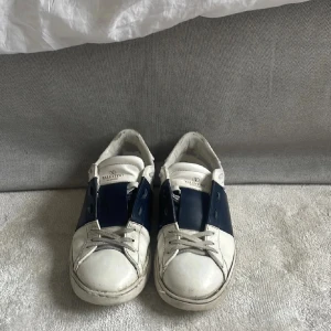 Valentino Open sneakers vit/marinblå - Snygga Valentino Open sneakers i vitt skinn med bred marinblå läderdetalj över mitten. Klassisk rund tå, platt sula och snörning framtill. Perfekta för dig som gillar exklusiva och stilrena sneakers med en twist.