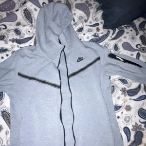 Äkta Nike Teech Fleece - Snygg grå zip hoodie från Nike med svart logga på bröstet, hel dragkedja och huva. Jackan har svarta detaljer längs bröstet och en praktisk ficka med dragkedja på ärmen. Använd endast ett fåtal gånger. Storlek S.