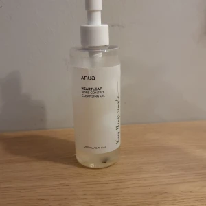 Anua Heartleaf Cleansing Oil 200ml - Anua Heartleaf Pore Control Cleansing Oil i genomskinlig plastflaska med pump, 200 ml. Oljan är lätt och genomskinlig, perfekt för att ta bort smink och rengöra porerna. Minimalistisk design med svart text och vit etikett. 