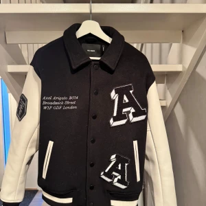 Svartvit varsity jacket Axel Arigato - Snygg svart och vit varsity jacket från Axel Arigato med stora broderade bokstäver och patch på ärmen. Jackan har kontrasterande vita ärmar i skinn och svart kropp i ull, samt coola detaljer med text och brodyr både fram och bak. Perfekt statement-plagg för höst och vinter.