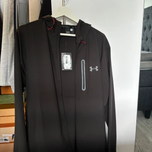 Svart vindjacka från Under Armour L - Svart vindjacka från Under Armour med huva och röda detaljer vid snörningen. Jackan har en bröstficka med dragkedja och Under Armour-logga på bröstet. Perfekt för blåsiga dagar och sportiga outfits.