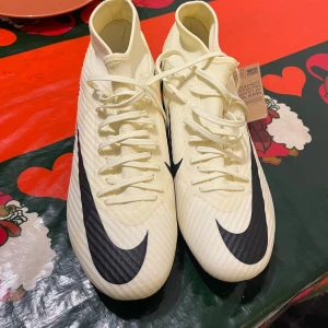 Nike vita fotbollsskor med svart swoosh - Snygga vita fotbollsskor från Nike med stor svart swoosh på sidan. Skorna har snörning och är tillverkade i syntetmaterial med en lätt struktur för bra bollkontroll. Perfekta för dig som vill sticka ut på planen med stilren design.