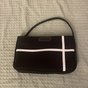 Svart axelväska från Lancôme - Snygg svart axelväska från Lancôme med raka former och stilrena vita detaljer i kors på framsidan. Väskan har ett kort axelband och dragkedja upptill. 