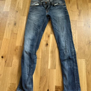 Blå jeans från Nudie Jeans, W30 L32 - Säljer ett par klassiska blå jeans från Nudie Jeans med snyggt slitna detaljer och orange sömmar. Modellen har raka ben, fem fickor och läderpatch med logga bak. Perfekta för dig som gillar en tidlös jeanslook.