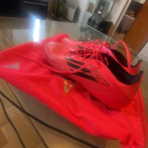 Röda Adidas F50 fotbollsskor - Snygga och lätta Adidas F50 fotbollsskor i en riktigt skarp röd färg med svarta detaljer. Skorna har snörning och platt sula med fasta dobbar för bra grepp på planen. Perfekta för dig som vill sticka ut på matchen och få maxad speed.