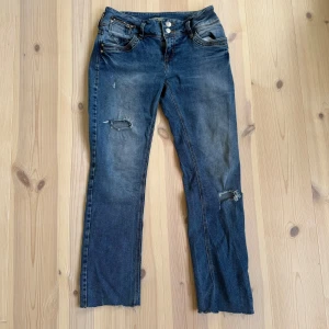 Blå jeans från LTB med slitningar - Säljer ett par blå jeans från LTB med snygga slitningar på benen och råa kanter nertill. Klassisk femficksmodell med dragkedja och dubbla knappar i midjan. Jeansen har kontrastsömmar och är i stretchig bomullsdenim.