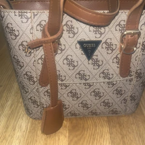 Beige handväska från Guess med monogram - Snygg beige handväska från Guess med bruna detaljer och klassiskt monogrammönster över hela väskan. Väskan har två handtag, justerbara remmar och dragkedja upptill. Perfekt storlek för det viktigaste och stilren design med Guess-logga i metall framtill.