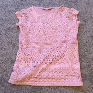 Rosa spetströja med kort ärm - Söt rosa blus i spets med korta ärmar och rund hals. Blusen har ett detaljerat spetsmönster med olika geometriska former och är lätt transparent. Perfekt för dig som gillar romantisk och feminin stil.