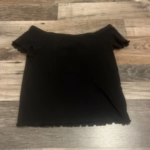 Svart offshoulder topp från KappAhl - Svart offshoulder topp från KappAhl med korta ärmar och vågiga kanter vid ärmslut och nederkant. Enkel och stilren design som passar perfekt till jeans eller kjol. Mjuk och stretchig känsla.