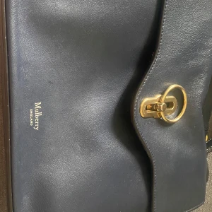 Mörkblå axelväska från Mulberry - Snygg mörkblå axelväska från Mulberry i slätt skinn med guldfärgade metalldetaljer och klassiskt lås framtill. Väskan har en stilren design med lock och präglad logga på framsidan. Perfekt för dig som gillar tidlös och lyxig stil.