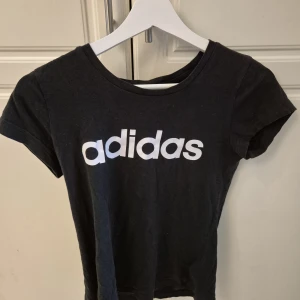 Svart Adidas t-shirt med logga - Svart t-shirt från Adidas med klassisk vit logga tryckt framtill. Modellen är kortärmad och har en rund halsringning. Tillverkad i mjuk bomull som är skön att ha på sig. Perfekt för dig som gillar sportig och enkel stil.