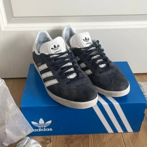 Adidas Gazelle mörkblå sneakers - Säljer ett par klassiska Adidas Gazelle sneakers i mörkblå mocka med vita detaljer och de ikoniska tre ränderna på sidorna med en ljusblå skolåda på köpet. Skorna har vit sula, snörning och Adidas-logga på plösen och hälen. Perfekta för dig som gillar retrostil och streetwear.