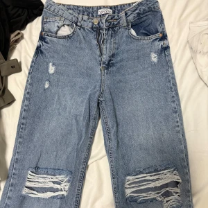 Blå ripped jeans från DeFacto - Säljer ett par ljusblå jeans från DeFacto med snygga slitningar och stora hål vid knäna. Jeansen har raka ben med fransade benslut. Perfekta för en avslappnad och trendig look.