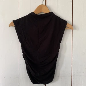 Svart croppad topp från Gina Tricot - Svart ärmlös croppad topp från Gina Tricot i mjuk bomull. Toppen har hög hals och snygg rynkning i sidorna som ger en cool siluett. Perfekt att styla med höga jeans eller kjol för en trendig look.