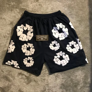 Mörkblå shorts från Denim Tears - Snygga mörkblå shorts från Denim Tears med unikt vitt blommigt mönster över hela plagget. De har elastisk midja och en bakficka. Perfekta för dig som vill sticka ut med streetwear-vibe och cool design.