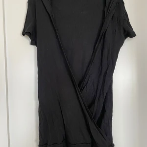 Svart omlott cardigan jacka med huva - Svart ribbad cardigan jacka från Lindex med omlottknäppning och huva. Jackan har korta ärmar och tre dekorativa knappar på sidan. Perfekt för lager-på-lager och chill vibes.