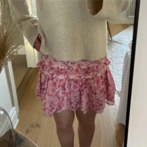 Rosa volangkjol med blommigt mönster - Supersöt kort kjol i rosa toner med blommigt mönster och volangdetaljer. Kjolen har smock i midjan för en bekväm passform och är gjord i ett lätt, luftigt material som passar perfekt till varmare dagar. Perfekt att styla med en stickad tröja eller en enkel topp.