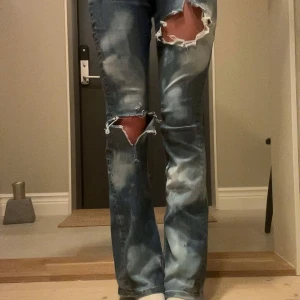 Blå slitna jeans med stora hål - Säljer ett par blå jeans med stora slitningar och hål över knäna. Jeansen har en boot cut fit och coolt blekta partier för en edgy look. Klassisk femficksmodell och låg midja. Perfekta för dig som gillar en avslappnad och trendig stil.