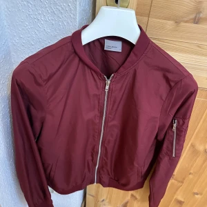 Vinröd bomberjacka från Vero Moda - Snygg vinröd bomberjacka från Vero Moda med dragkedja framtill och klassisk krage. Jackan har lång ärm och en extra ficka med dragkedja på ena ärmen. 