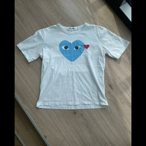 Vit Comme des Garçons Play t-shirt - Vit t-shirt från Comme des Garçons Play med ett stort blått hjärta med ögon och ett litet rött hjärta på bröstet. Klassisk passform, rund hals och korta ärmar. Tillverkad i mjuk bomull, perfekt för en clean och cool look. Storlek M och bara använd en gång. Har kvar påsen också.