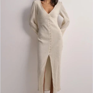 Beige långklänning med v-ringning - Stilren beige långklänning från Nelly med v-ringad hals, långa ärmar och knappar framtill. Klänningen har en slits framtill och är tillverkad i ett ribbat material som ger en snygg struktur.