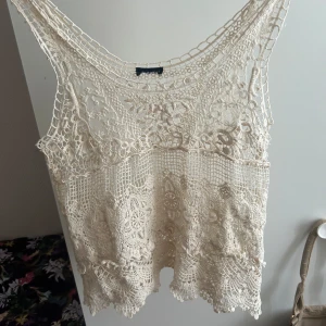 Beige spetsig linnetopp med virkad look - Supersöt beige linnetopp med detaljerad virkad spets och blommiga mönster. Toppen är ärmlös och har en luftig, bohemisk känsla med fina kanter nertill. Perfekt för varma dagar och ger en romantisk vibe till din outfit.
