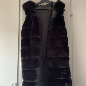 Svart lång pälsväst med huva - Säljer en svart lång pälsväst från Fashion med breda, horisontella paneler och stor huva. Västen är fluffig och har en lyxig känsla, perfekt för att ge din outfit extra edge. Fodrad insida för extra komfort.